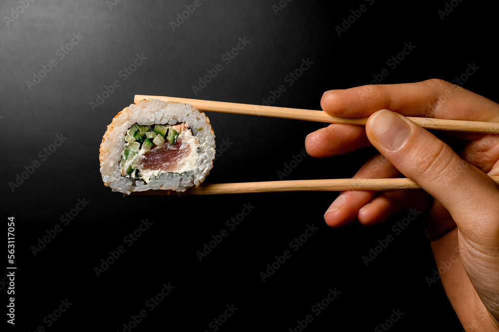 side view of hand holding one piece of roll california with salmon garnished sesame seeds with bamboo chopsticks