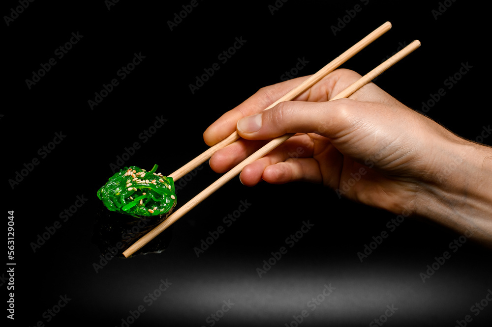 hand holds roll of gunkan sushi with chukka and sesame seeds wrapped in nori seaweed with chopsticks on dark background.