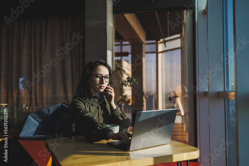 Young freelancer woman. Young woman working with laptop in coworking space and talking on phone, sitting in open space with cup of coffee and smiling while holding glasses.