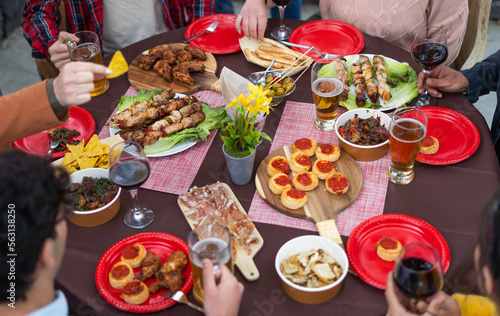 A beautifully set table featuring an array of delicious grilled meats, mini pizzas, crispy fries alcoholic beverages. Multiracial group of friends having summer party outdoor