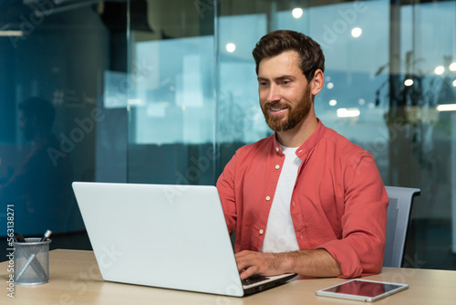 Successful businessman in red shirt happily working with laptop inside office, mature man with beard at workplace typing on keyboard smiling satisfied with work results and achievement.