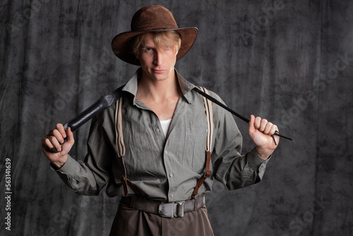treasure hunter, a hero of a retro-style adventure. A young man in a hat and breeches with suspenders with a whip in his hands. Posing in the studio on a gray background