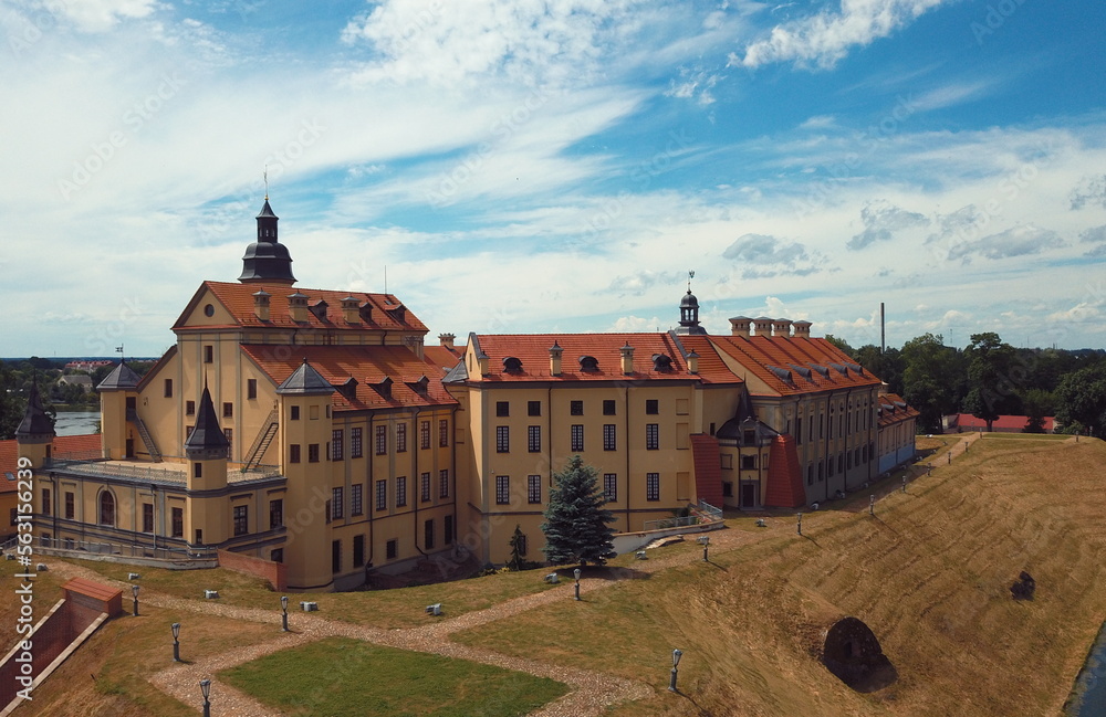 Medieval Eastern European castle with dark yellow and orange brick walls stands on a hill with a defensive earthen rampart to protect against a siege on a blue summer background