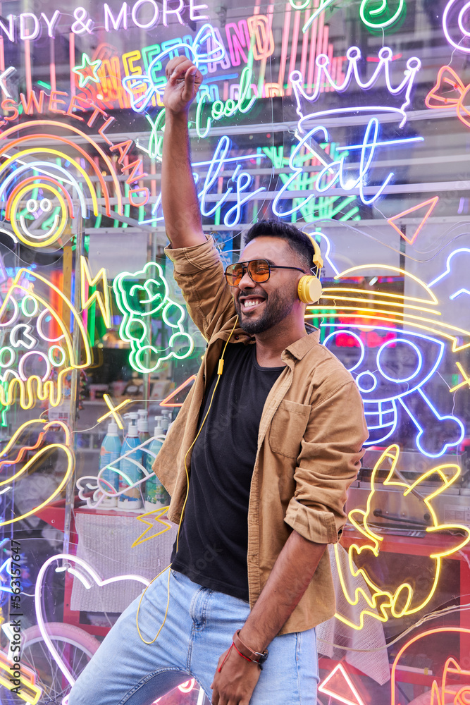 Hispanic male listening to music with headphones. young man with beard, sunglasses and casual clothes dancing with neon lights background.