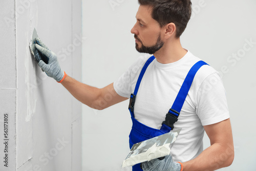 Professional worker in uniform plastering wall with putty knife indoors photo