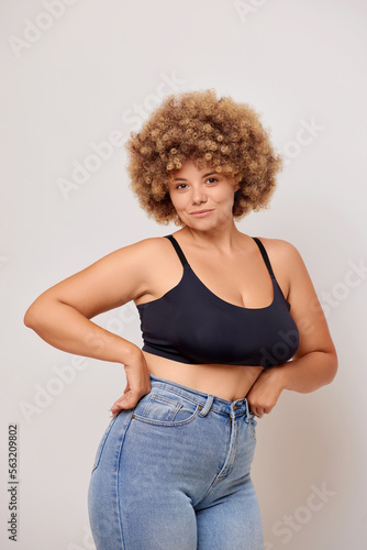 Confident plus size woman with Afro hair feels confident, wears jeans and black lingerie bra, poses in studio over white wall 