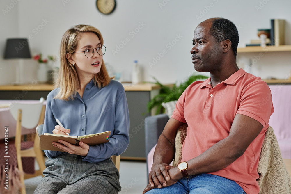 Young psychologist asking questions to patient and making notes in notepad during psychotherapy session Young psychologist asking questions to patient and making notes in notepad during psychotherapy session