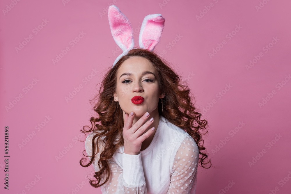 beautiful young woman with red long hair posing in a white sweater and rabbit ears. Happy easter concept. Looking into the camera, pink background. beautiful young woman with red long hair posing in a white sweater and rabbit ears. Happy easter concept. Looking into the camera, pink background.