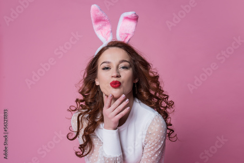 beautiful young woman with red long hair posing in a white sweater and rabbit ears. Happy easter concept. Looking into the camera, pink background.