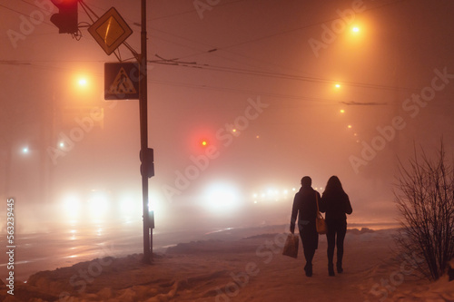 City at night in dense fog. People walking down a city street in the fog. A cityscape at night. Fog in the city, evening. Women walking in the dark in the light of car headlights. 