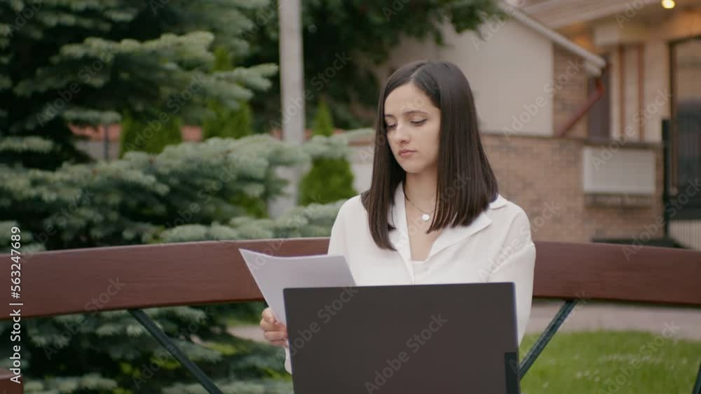 Woman making comparison of papers and laptop version of it outside office. large view of brunette working remolety from park. Woman making comparison of papers and laptop version of it outside office. large view of brunette working remolety from park.