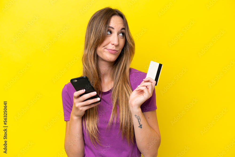 Young caucasian woman isolated on yellow background buying with the mobile with a credit card while thinking