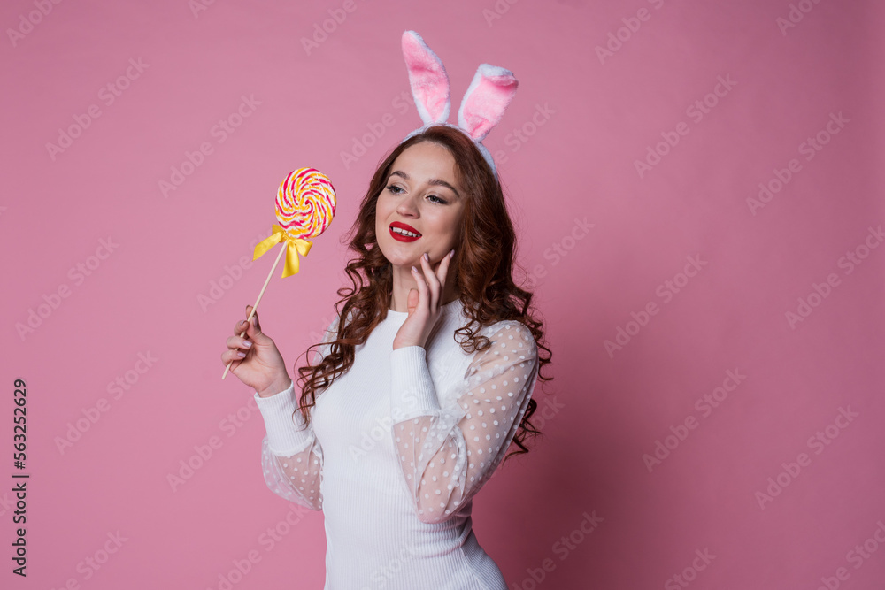 happy easter, beautiful young girl with long red hair in white easter bunny ears holding lollipop on pink background