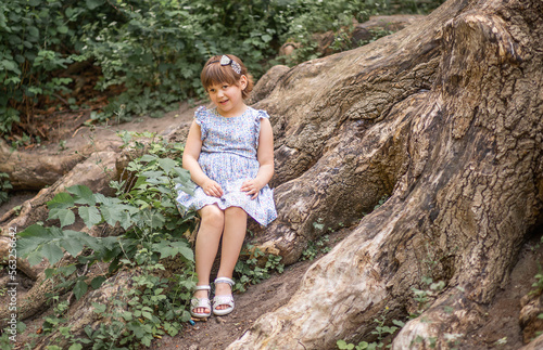 A cute little girl sits on huge tree roots, walks in the fresh air in summer, the child is wearing a summer dress. Summer sunny day. Children's holidays.
