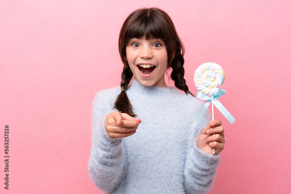 Little caucasian girl holding a lollipop surprised and pointing front Little caucasian girl holding a lollipop surprised and pointing front