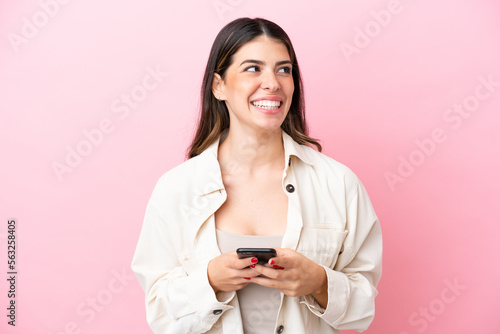 Young Italian woman isolated on pink background using mobile phone and looking up
