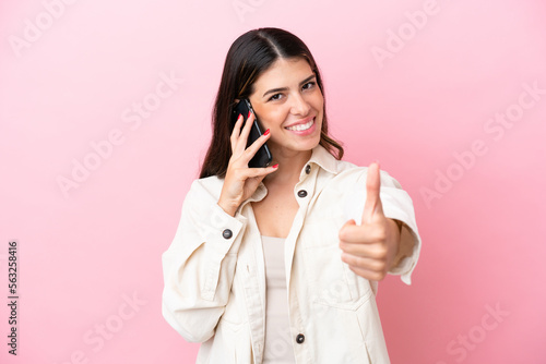 Young Italian woman isolated on pink background keeping a conversation with the mobile while doing thumbs up