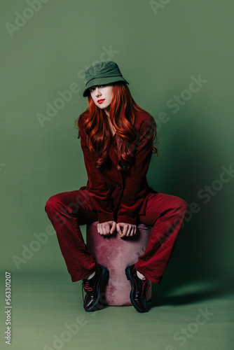 Stylish woman in sunglasses, hat and red suit sits on green background photo