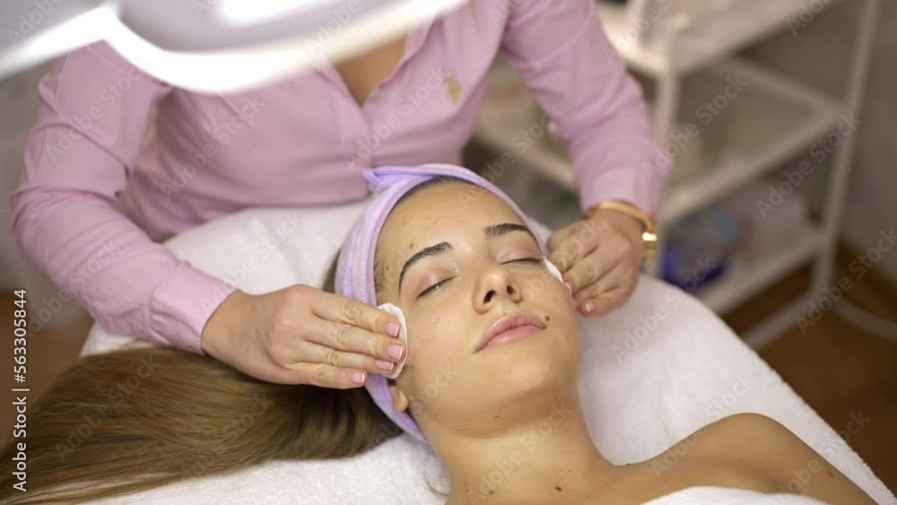 Close-up shot of beautician wipes the face of a beautiful girl with cotton pads under the lamp light. European young woman in a beauty salon. Skin care and wrinkle treatment Close-up shot of beautician wipes the face of a beautiful girl with cotton pads under the lamp light. European young woman in a beauty salon. Skin care and wrinkle treatment