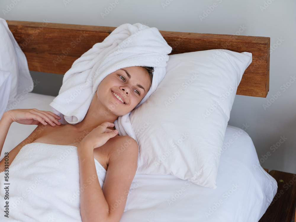 A woman lies on the bed after a shower with wet hair with a towel on her head in the bedroom and strokes her body skin, a cute smile with teeth and weekend rest in bed, women's beauty day, lifestyle A woman lies on the bed after a shower with wet hair with a towel on her head in the bedroom and strokes her body skin, a cute smile with teeth and weekend rest in bed, women's beauty day, lifestyle