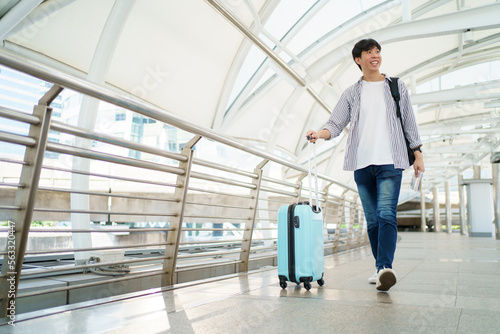 Happy cheerful Asian handsome young man walking in the city railway station alone with suitcase. 