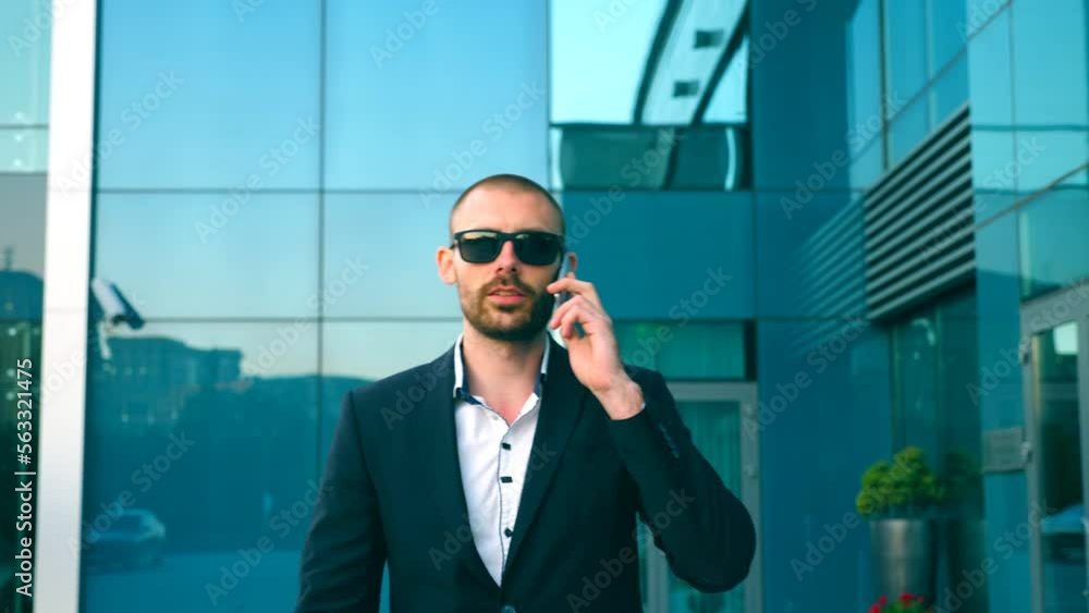 Portrait of businessman in sunglasses talking on phone and walking in street. Young man having business conversation during commuting to work. Confident guy in suit being on his way to office building Portrait of businessman in sunglasses talking on phone and walking in street. Young man having business conversation during commuting to work. Confident guy in suit being on his way to office building