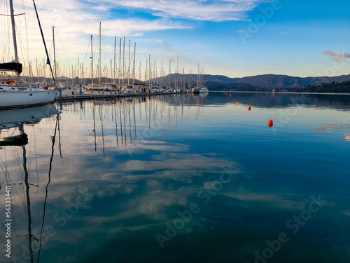 Fototapeta Naklejka Na Ścianę i Meble -  Sail boat yacht marina port harbour with calm blue ocean sea reflecting sky clouds ship masts mountains. and mooring buoys. With nobody and copy space.