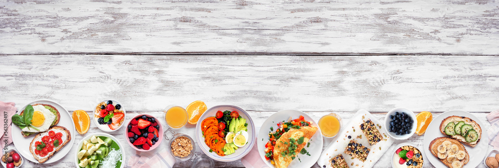 Healthy breakfast food bottom border. Top down view over a white wood banner background. Table scene with omelette, nutritious bowl, toasts, granola bars, smoothie bowl, yogurts, fruit. Copy space.