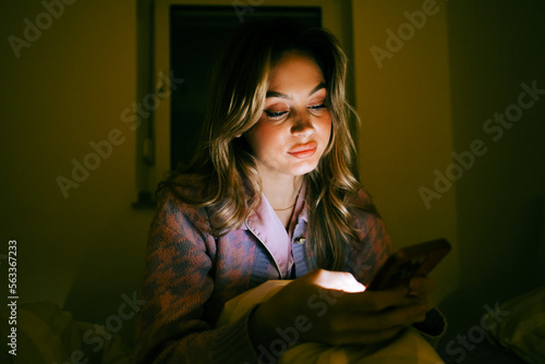 Young caucasian woman using smartphone, sitting on a bed at home in the evening.