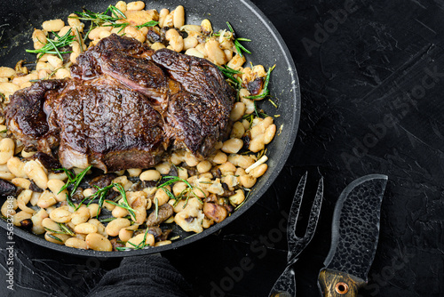 Legendary grilled rib eye with beans, on frying cast iron pan, with meat knife and fork, on black stone background, with copy space for text