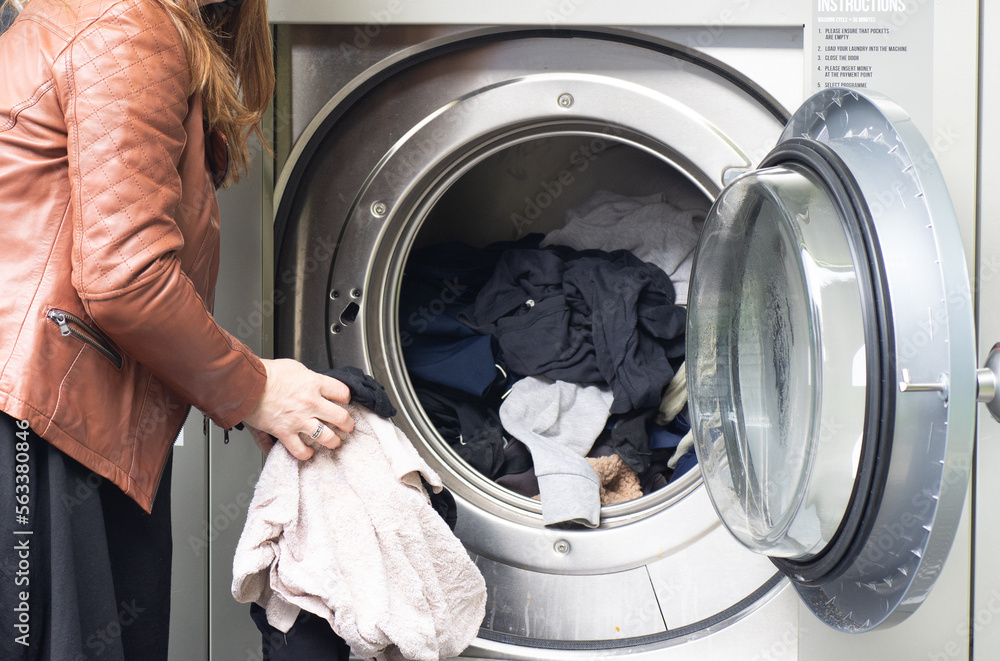 middle-aged woman in black dress and brown jacket, using self-service laundromat, introducing dirty clothes and using card payment, horizontal image, copy space