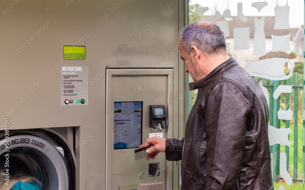 senior male, wearing jeans and brown jacket, using self-service laundry, inserting dirty clothes and using card payment, horizontal image, copy space senior male, wearing jeans and brown jacket, using self-service laundry, inserting dirty clothes and using card payment, horizontal image, copy space
