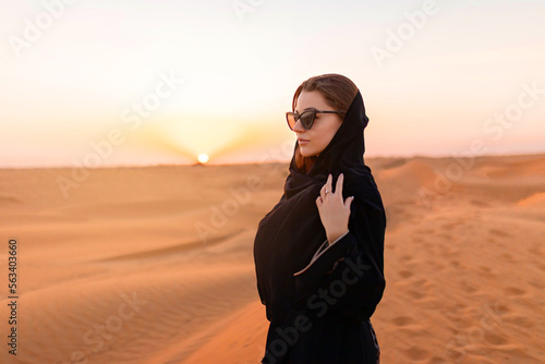 Beautiful mysterious woman in traditional arabic black long dress stands in the desert on sunset