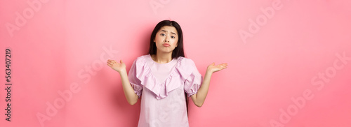 Sorry cant help. Clueless asian girl shrugging with sad face, know nothing, standing indecisive against pink background photo