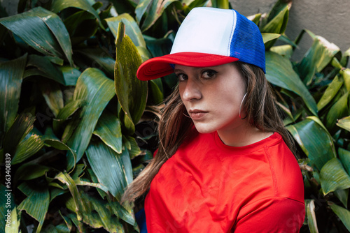 Young woman with plants on background. Serious face. Teenage girl wearing vibrant color clothing. Female model wearing red tshirt. Red and blue. Young caucasian female model.