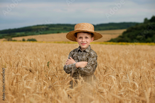 A smiling little farmer boy in a plaid shirt and straw hat poses for a photo in a wheat field. Heir of farmers