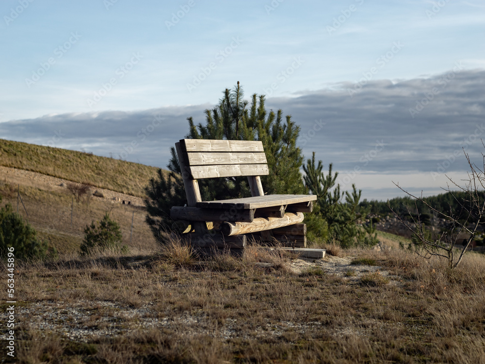 Naklejka premium Empty park bench on top of a hill in the nature. The calm and idyllic landscape is in the background. Nice place to rest in a rural area without people.