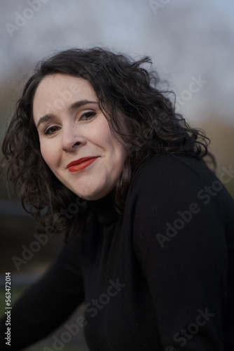A woman with pale skin and black, wavy hair poses confidently outdoors.