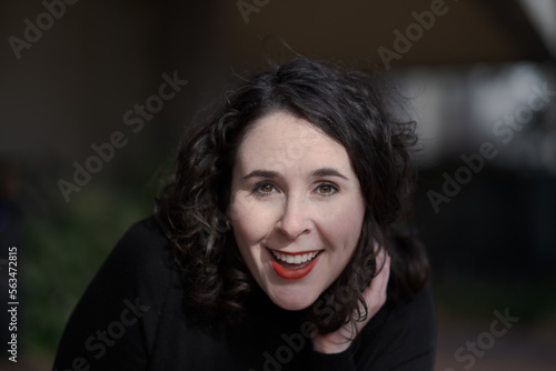 A woman with pale skin and black, wavy hair poses confidently outdoors.