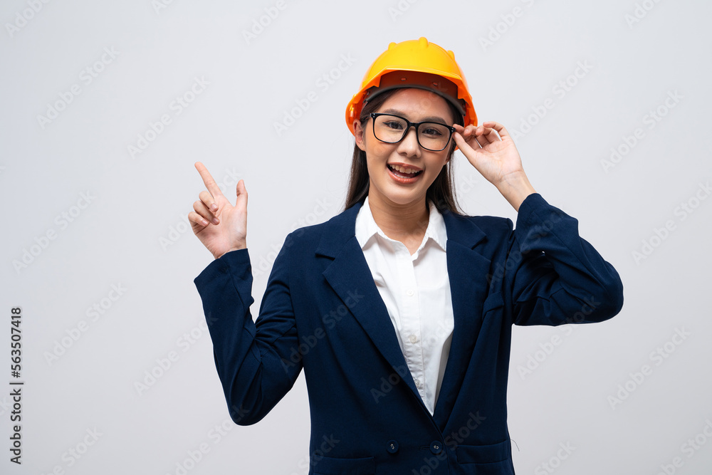 Portrait of Asian female engineer with hard hat pointing at copy space isolated on grey background.