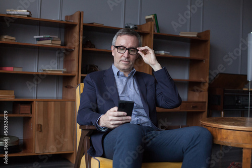 Attractive elderly grey haired man in glasses working on mobile phone, sitting on chair in office