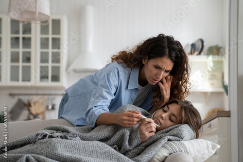 Worried mother checking measuring temperature of sick child daughter lying under blanket at home, anxious mom looking at thermometer, taking care about unwell ill kid with high fever resting in bed
