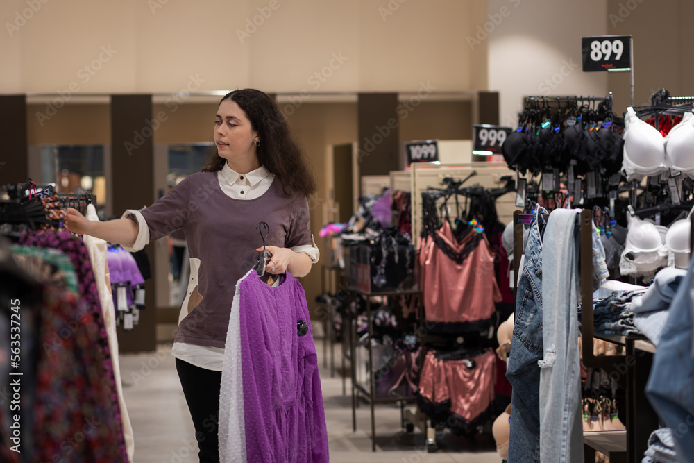 Young Caucasian woman chooses clothes in store. In background there are fitting rooms and hangers with clothes. Concept of shopping and purchase Young Caucasian woman chooses clothes in store. In background there are fitting rooms and hangers with clothes. Concept of shopping and purchase