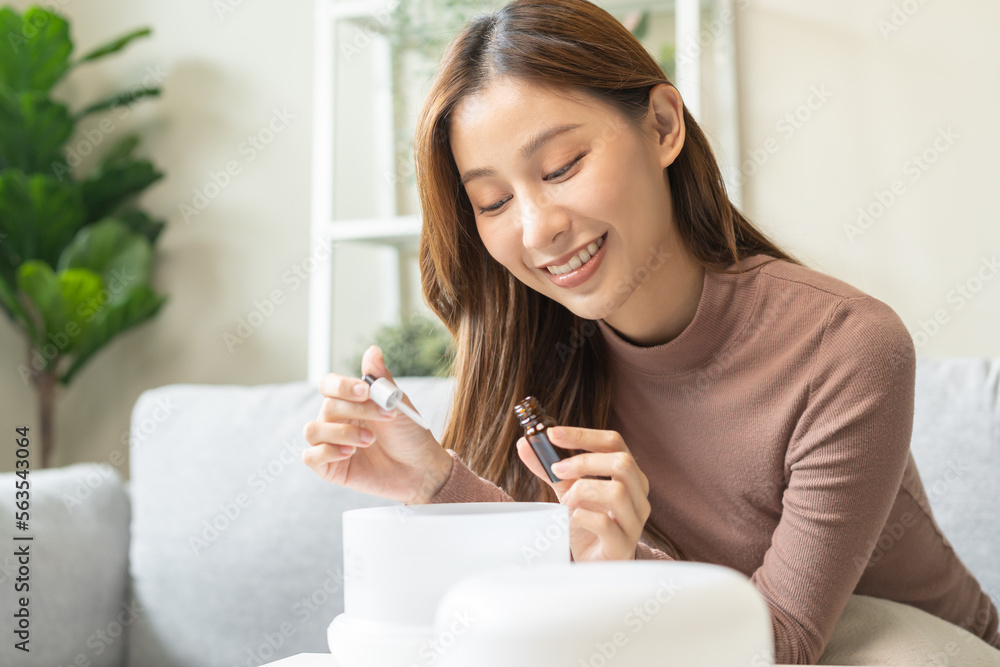 Happy asian young woman, girl hand holding bottle of essential and drop oil into air humidifier, enjoy aromatherapy steam scent diffuser comfortable while rest in living room, apartment at home. Happy asian young woman, girl hand holding bottle of essential and drop oil into air humidifier, enjoy aromatherapy steam scent diffuser comfortable while rest in living room, apartment at home.