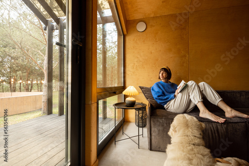 Woman reads a book while sitting relaxed on a couch by the window with her dog at wooden cottage in forest. Concept of home comfort and rest in a house on nature. Wide angle view