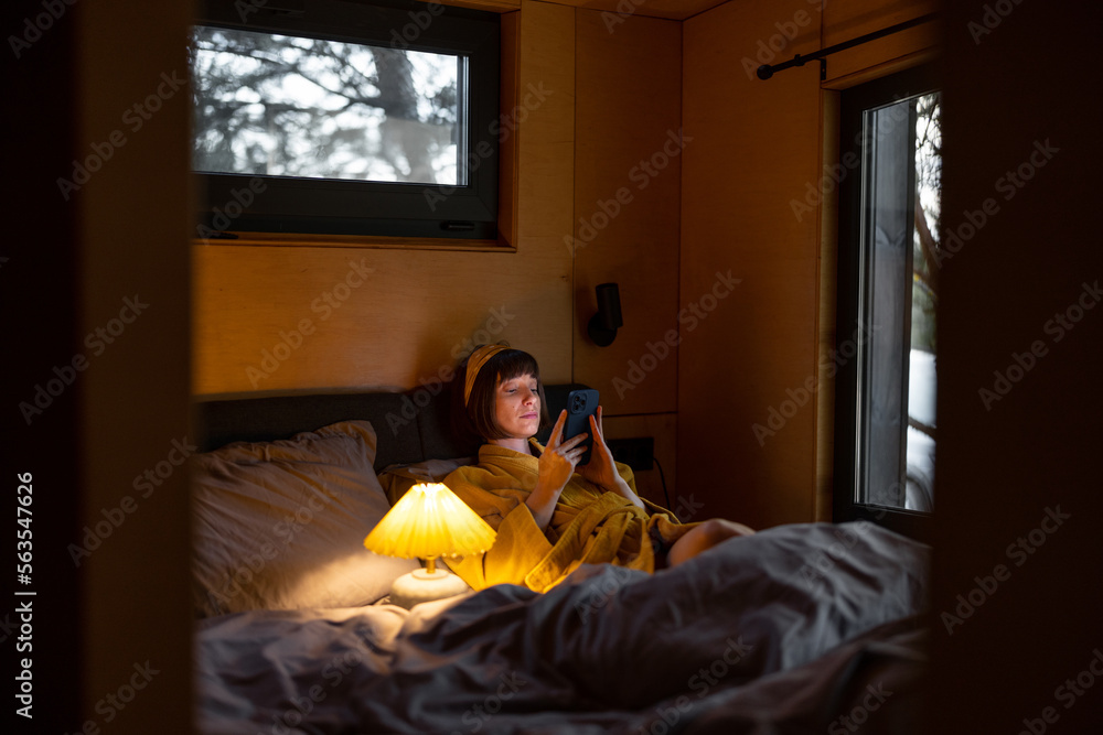 Naklejka premium Woman in yellow bathrobe uses a smartphone while lying in bed at tiny bedroom of wooden cabin on nature. Communication online during bedtime and addiction to smartphone