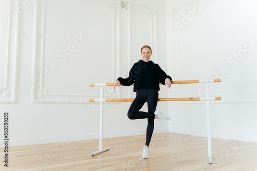Foto Positive slim woman has dance class, practices in studio near ballet barre, stands on one leg, wears black outfit and white sportshoes, has strong muscles, nice body shape