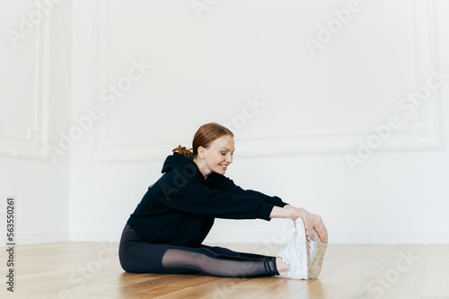 Indoor shot of smiling redhead woman stretches legs, focused into distance, touches feet, wears white sportshoes, has gymnastic exercises, prepares for cardio training. Healthy lifestyle concept