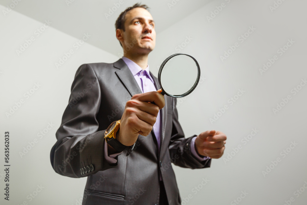 Serious hipster business man in suit holding magnifying glass and looking at camera, over grey background Serious hipster business man in suit holding magnifying glass and looking at camera, over grey background