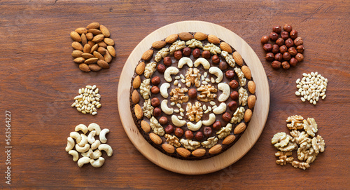 Beautiful homemade festive chocolate cake decorated with various types of nuts on wooden table. Walnuts, hazelnuts, cashews, pine nuts and almonds next to it. Flat lay, close-up, top view, mock up